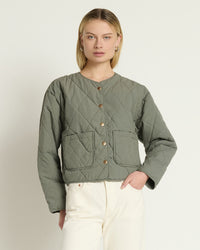 A woman wearing the Aspen Quilted Jacket in Castor Grey by Nation LTD and off-white pants stands against a plain light background.