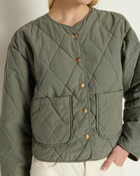 A person wears the Aspen Quilted Jacket in Castor Grey by Nation LTD, featuring gold snap buttons and two front pockets, paired with off-white pants. The photo is cropped at the shoulders and hips.