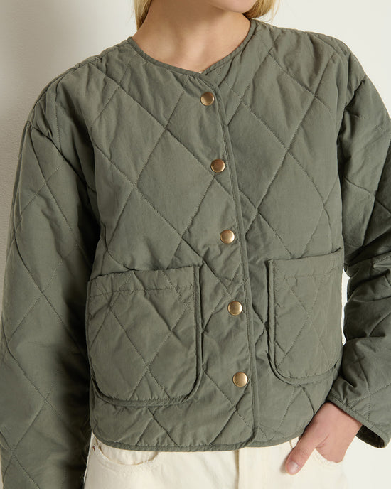 A person wears the Aspen Quilted Jacket in Castor Grey by Nation LTD, featuring gold snap buttons and two front pockets, paired with off-white pants. The photo is cropped at the shoulders and hips.