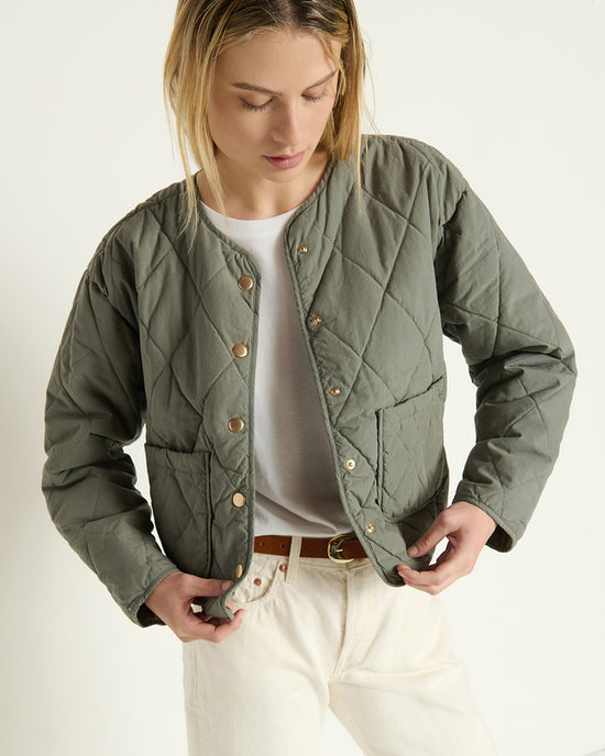 A person wearing the Nation LTD Aspen Quilted Jacket in Castor Grey over a white shirt and off-white pants stands against a plain background, looking down while adjusting the front snap buttons.