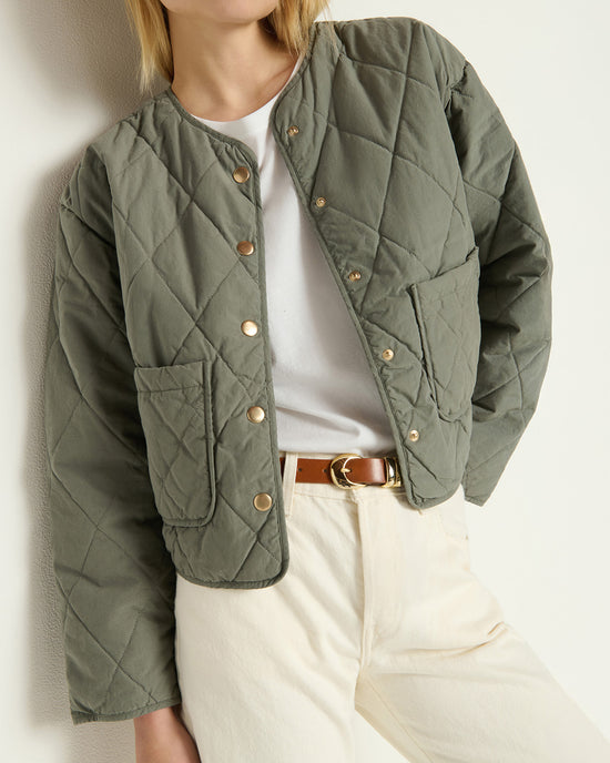 A person wears the Nation LTD Aspen Quilted Jacket in Castor Grey, styled with a white t-shirt, cream pants, and a brown belt, posing with one hand in their pocket against a plain light background.