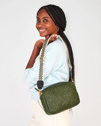 Woman with braided hair smiling over her shoulder, holding a green Clare V. bag with a Checker Adj Crossbody Strap in Black & Cream, wearing a light blue blouse.