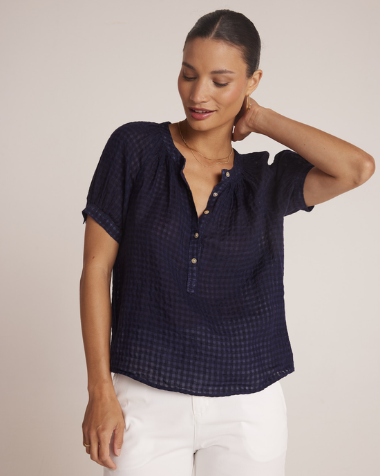 A woman wearing a Bella Dahl Short Sleeve Raglan Pullover in Tropic Navy and white pants, gently touching her neck and smiling subtly against a neutral background.