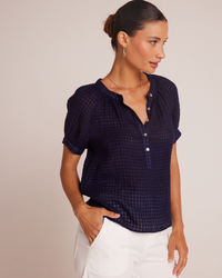 Woman in a dark blue Bella Dahl Short Sleeve Raglan Pullover in Tropic Navy henley button down and white pants, looking to the side, against a neutral background.