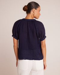 A woman wearing a navy blue Bella Dahl Short Sleeve Raglan Pullover in Tropic Navy and white pants, viewed from the back with a focus on the blouse's textured detail. She has her hair in a bun and wears
