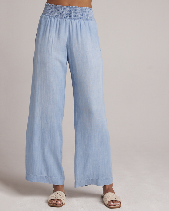 A person standing in light blue Bella Dahl Smocked Waist Wide Leg Pant in Caribbean Wash trousers and brown sandals against a neutral background.