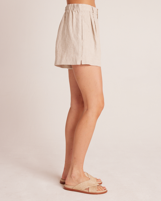 A person wearing Bella Dahl's Pleat Front Trouser Short in Linen Sand with an elastic waistband and tan sandals stands against a pale background, displaying a side view from the waist down.