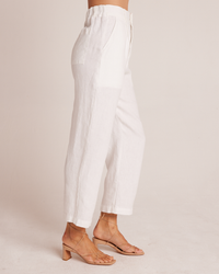 A person wearing Bella Dahl's Relaxed Pleat Front Trouser in White and tan strappy sandals, shown from the waist down, standing against a neutral background.