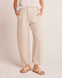 A person wearing Bella Dahl's Relaxed Pleat Front Trouser in Linen Sand and a white shirt, paired with beige sandals, standing against a neutral background.
