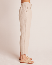 A person wearing Bella Dahl's Relaxed Pleat Front Trouser in Linen Sand and beige sandals stands against a neutral background. Only the lower half of the body is visible.