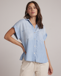 A woman wearing a Bella Dahl Two Pocket S/S Shirt in Caribbean Wash button-down shirt and wide leg jeans, standing with one hand on her hip.
