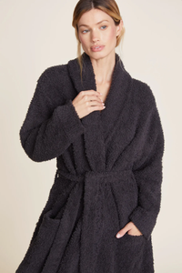 A woman in a dark Barefoot Dreams Cozychic Solid Robe in Carbon with a tie waist standing against a neutral background.