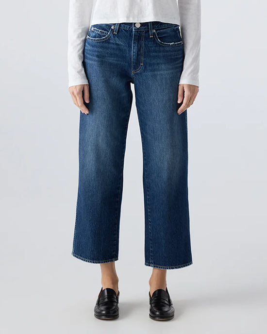 A person wearing AMO's Billie in Metaphor mid rise straight leg cropped blue jeans with black loafers and a white long-sleeve shirt, standing against a plain light background.