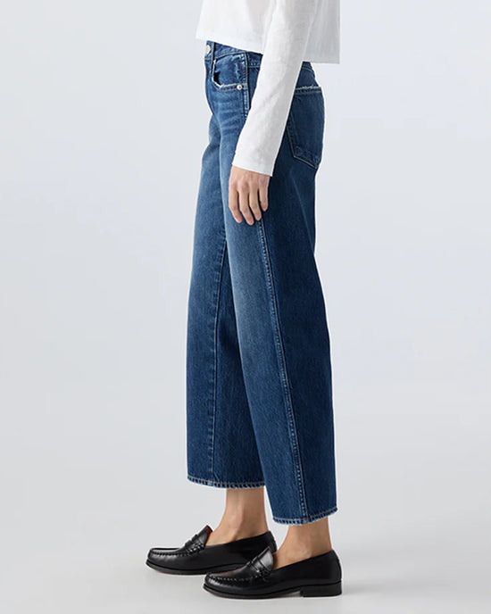 A person in AMO’s Billie in Metaphor jeans, a white long-sleeve top, and black loafers stands sideways against a plain light background.