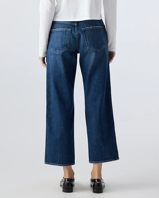 A person wearing AMO's Billie in Metaphor dark blue mid rise straight leg jeans and a white long-sleeve top is standing with their back to the camera. The jeans have large back pockets, paired with black shoes.