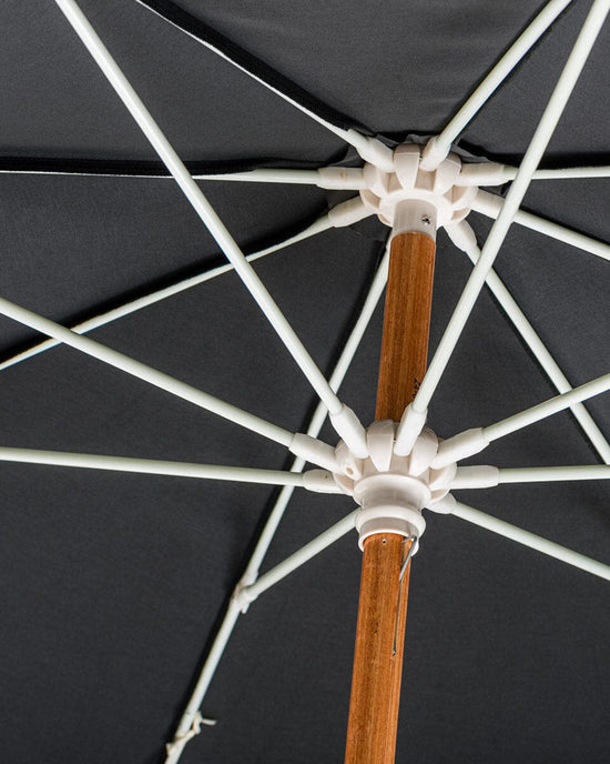 Close-up of the Amalfi Umbrella - Rivie Black by Business & Pleasure Co, highlighting its wooden pole, white metal support ribs, and Mediterranean-inspired silhouette.