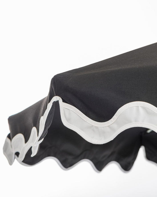 A close-up of the Business & Pleasure Co Amalfi Umbrella - Rivie Black, featuring Mediterranean style, UV and water-resistant black fabric with a white scalloped trim, shown against a plain white background.