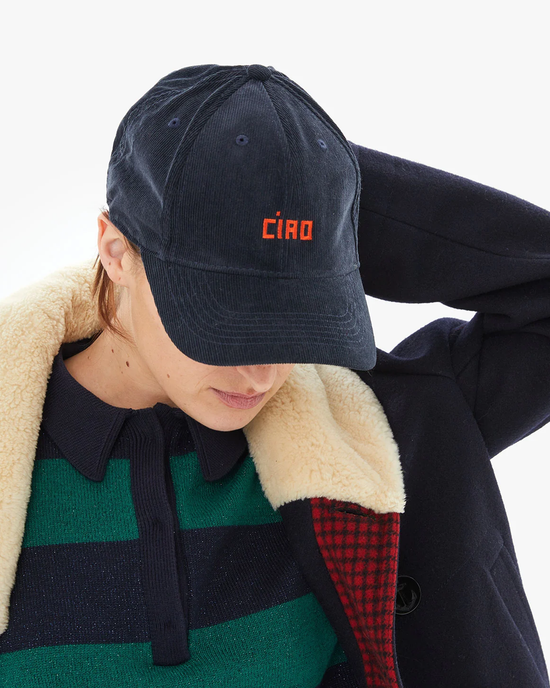Person wearing a Clare V. Emb Petit Block Ciao Corduroy Baseball Hat in Navy w/ Bright Poppy and a coat with a shearling collar.
