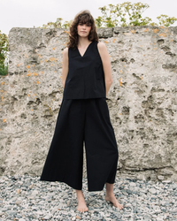Person in Mazu Pants in Black and a black sleeveless top made from organic cotton, standing barefoot on a pebble ground with a concrete wall in the background. The outfit is from Beaumont Organic.