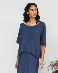A person is wearing the loose-fitting Alessia-Leigh Top in Denim Blue, made from recycled cotton, paired with a matching skirt. They stand against a neutral background with flowers on the side, showcasing Beaumont Organics commitment to ethical fashion.