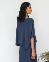A person with long, dark hair is wearing a Beaumont Organic Alessia-Leigh Top in Denim Blue and a loose skirt, standing sideways against a plain background with flowers at the bottom.
