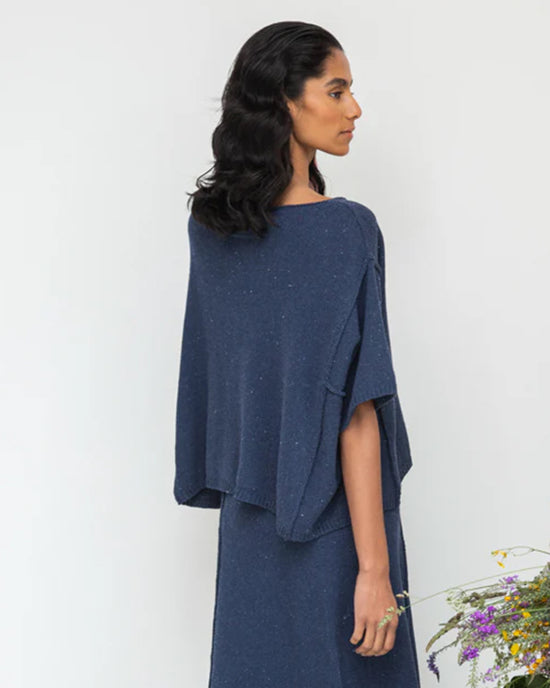 A person with long, dark hair is wearing a Beaumont Organic Alessia-Leigh Top in Denim Blue and a loose skirt, standing sideways against a plain background with flowers at the bottom.