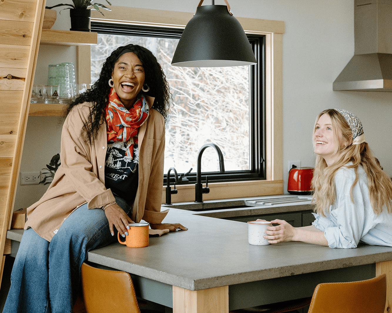 Two women smiling and chatting in a modern kitchen, one sitting on the counter holding a mug, the other sitting at the counter with a mug. Natural light comes through a window behind them.