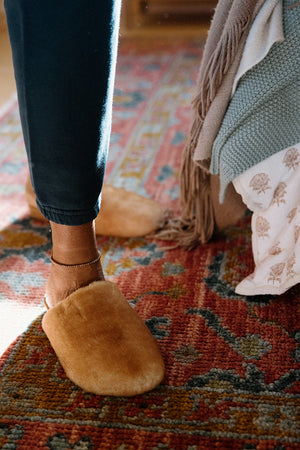 Person at home wearing comfortable slippers and resting one foot on a patterned rug.
