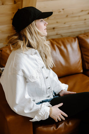Woman sitting on a brown sofa wearing a black cap and white denim jacket.