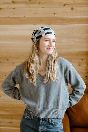 A smiling young woman in a gray sweater and striped headband stands with her hands on her hips against a wooden wall background.