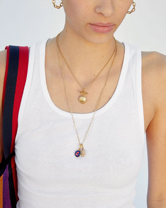 A person wears a white ribbed tank top, gold hoop earrings, and two layered gold necklaces: a Clare V. Fine Paperclip Charm Chain 24" in 14K Gold Vermeil and another with a beaded hand-shaped charm and small round blue pendant.