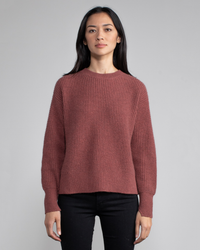 Woman in a terracotta Margaret O'Leary Nora Pullover in Cinnamon sweater and black pants standing against a grey background.