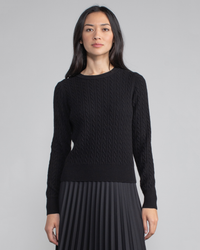 A woman in a Margaret O'Leary Baby Cable Pullover in Black and pleated skirt standing against a gray background.