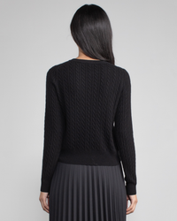 A woman standing with her back to the camera, wearing a black Margaret O'Leary Baby Cable Pullover and a black pleated skirt.