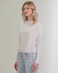 A woman in a light grey Margaret O'Leary Cashmere Ballet Wrap in Mist and blue jeans standing against a neutral background.