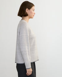A woman with short brown hair stands in profile against a plain light background, wearing the Margaret O'Leary Mendocino Pullover in Fog Combo and dark trousers.