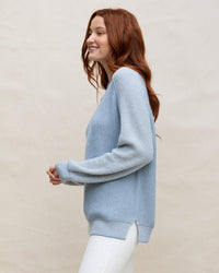 A woman with long red hair wears the Margaret O'Leary Baseball Tunic in Denim, paired with white pants, standing in profile and smiling against a plain light beige background.
