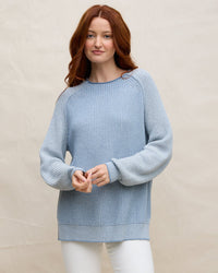 A woman with long red hair wears the Margaret O'Leary Baseball Tunic in Denim and white pants, standing against a plain beige background and looking at the camera with a gentle smile.
