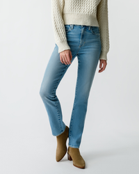 A person wearing light blue AMO Chloe Long Length in Liason slim fit jeans, a white knit sweater, and brown ankle boots standing against a plain background.