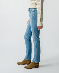 Person standing sideways wearing AMO Chloe Long Length in Liason high rise blue jeans and brown boots with a white knit sweater.