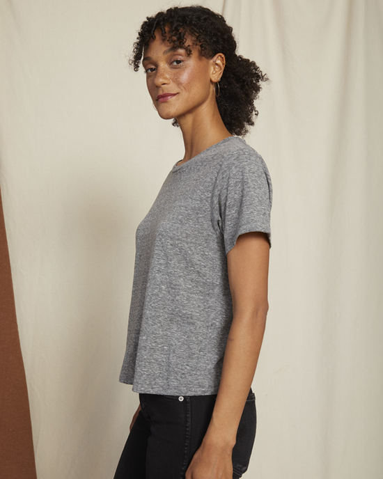 A woman in an AMO Classic Tee in Heather Grey with distressed edges and black pants standing against a beige background, looking over her shoulder with a slight smile.
