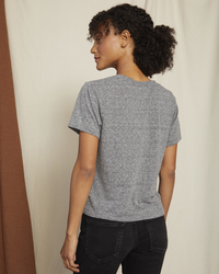 A woman in an AMO Classic Tee in Heather Grey with distressed edges and black jeans standing with her back to the camera, looking over her shoulder.