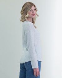 Woman in a Margaret O'Leary Shirttail Easy Boatneck in Ivory/Salt and jeans standing against a plain background, smiling over her shoulder.