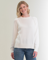 Woman smiling in a Margaret O'Leary Livia Pullover in Ivory, a 100% cotton sweater and blue jeans standing against a neutral background.