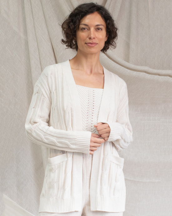 A woman in a white, oversized Margaret O’Leary Nina Cable Cardi in Oat standing in front of a draped cloth background.