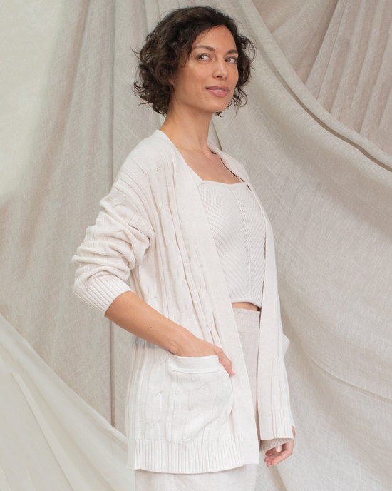 A woman in a Margaret O'Leary Nina Cable Cardi in Oat standing confidently against a draped fabric background.