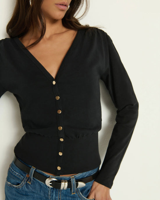A woman wears the Colette Cardigan Top in Jet Black by Nation LTD, made from soft Peruvian cotton with gold buttons, styled with blue jeans and a black belt with a silver buckle. Her head is slightly turned and her hair falls loosely.