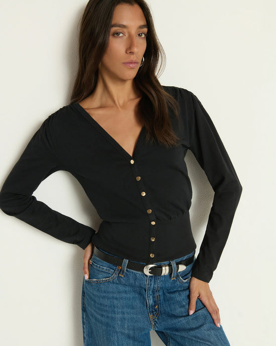 A woman with long brown hair poses confidently in the Nation LTD Colette Cardigan Top in Jet Black, a buttoned V-neck long-sleeve made from Peruvian cotton, paired with blue jeans and a black belt against an off-white wall.