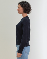 Side profile of a woman with curly hair wearing a dark Margaret O'Leary Lola Pullover in Navy and jeans against a neutral background.