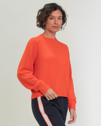 Woman in an organic cotton Margaret O'Leary Lola Pullover in Tomato, orange sweater and black trousers with a red stripe.